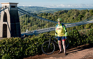 A cyclist fundraising for Anti-Slavery International stands in front of a bridge across a beautiful landscape. Credit James Markham.