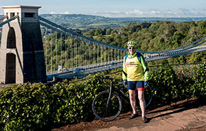 A cyclist fundraising for Anti-Slavery International stands in front of a bridge across a beautiful landscape