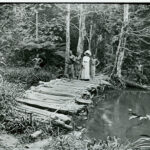 Black and white photo showing a white woman in a full dress and hat with several black people crossing a log bridge over a river