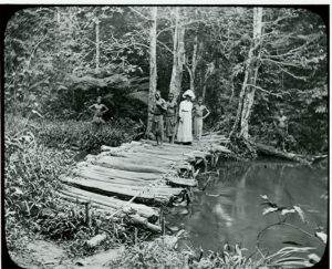 Black and white photo showing a white woman in a full dress and hat with several black people crossing a log bridge over a river