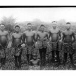 Black and white photo showing a group of semi-clad men holding baskets for rubber collection
