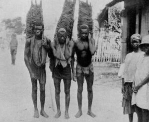 Black and white photo showing three enslaved black men carrying heavy burdens