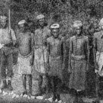 Black and white photo showing a group of black slaves with shackles and chains standing in a line, guarded by an askari or soldier