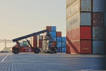 A shipping container is being unloaded from a truck at a port terminal.