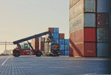 A shipping container is being unloaded from a truck at a port terminal.